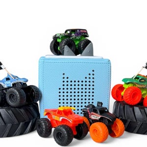 Puede incluir: Una colección de camiones monstruo de juguete en varios colores, incluyendo rojo, verde y negro, con neumáticos de gran tamaño. Algunos camiones tienen banderas a cuadros. Un altavoz azul claro en forma de cubo está en el fondo.