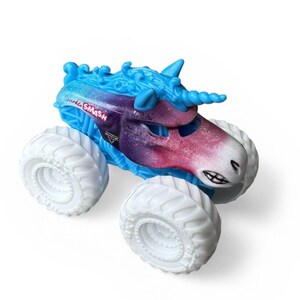 K&ouml;nnte beinhalten: Ein Spielzeug-Monstertruck mit Einhorn-Design. Der Truck hat eine blau-violette Karosserie mit wei&szlig;en R&auml;dern. Der Text "Shark SMASH" ist auf der Seite des Trucks zu sehen.