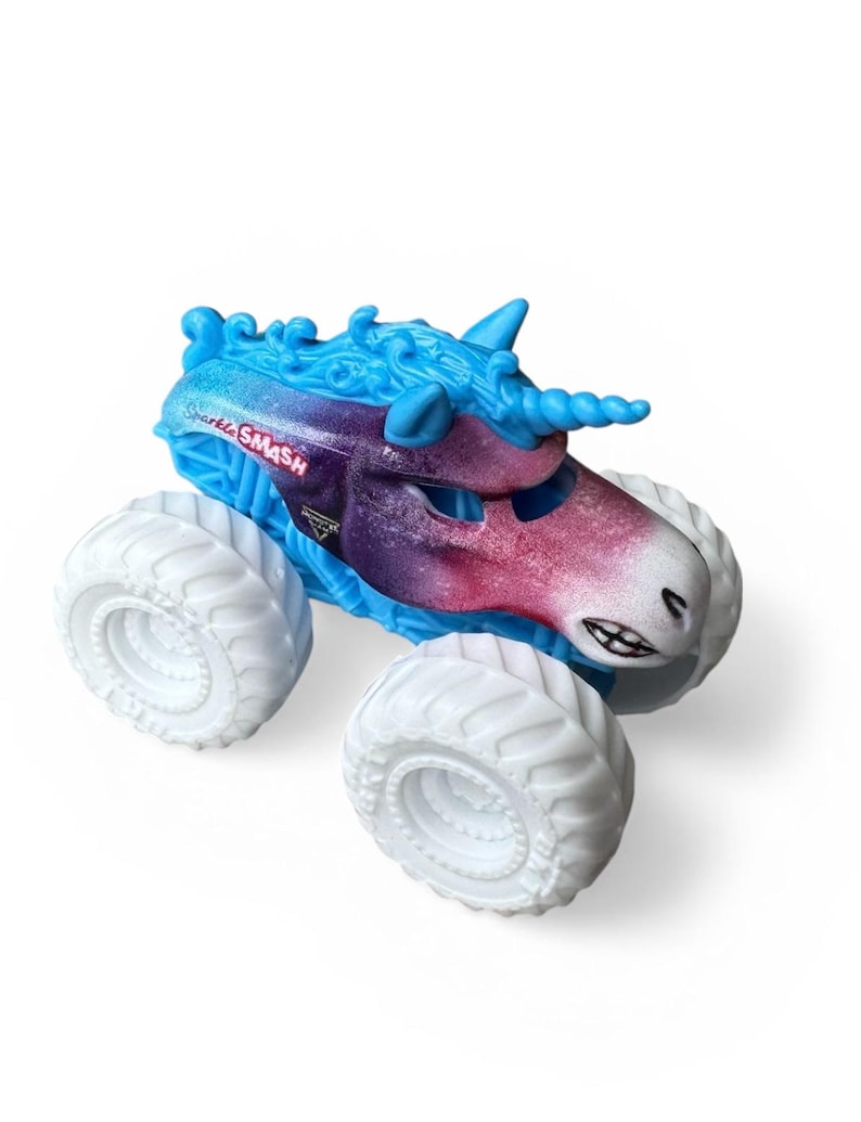 K&ouml;nnte beinhalten: Ein Spielzeug-Monstertruck mit Einhorn-Design. Der Truck hat eine blau-violette Karosserie mit wei&szlig;en R&auml;dern. Der Text "Shark SMASH" ist auf der Seite des Trucks zu sehen.