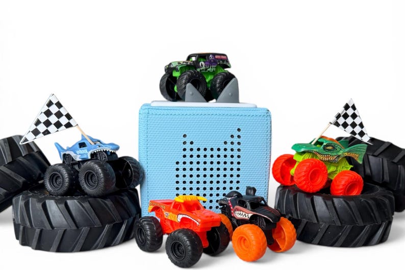 K&ouml;nnte beinhalten: Eine Sammlung von Spielzeug-Monstertrucks in verschiedenen Farben, darunter Rot, Gr&uuml;n und Schwarz, mit &uuml;bergro&szlig;en Reifen. Einige Trucks haben karierte Flaggen. Im Hintergrund befindet sich ein hellblauer, w&uuml;rfelf&ouml;rmiger Lautsprecher.