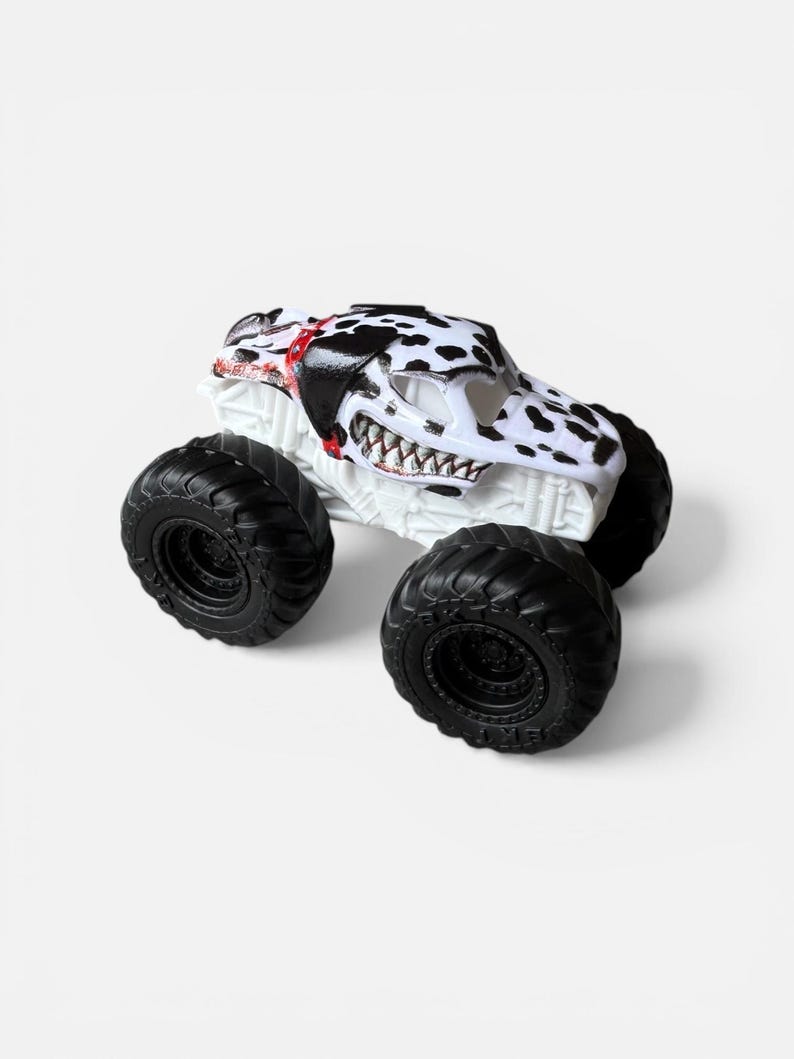 K&ouml;nnte beinhalten: Ein wei&szlig;es Monstertruck-Spielzeug mit schwarz-wei&szlig;en Dalmatiner-Punkten auf der Kabine. Der Truck hat gro&szlig;e schwarze Reifen und einen detaillierten, zahnartigen K&uuml;hlergrill. Die Kabine hat rote Akzente.