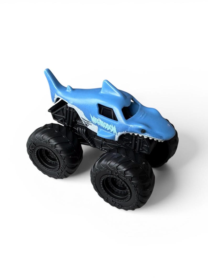 K&ouml;nnte beinhalten: Ein blaues Spielzeug-Monstertruck mit einem haif&ouml;rmigen K&ouml;rper. Der Truck hat gro&szlig;e schwarze Reifen und das Wort "MEGASHARK" ist auf der Seite aufgedruckt. Der Haik&ouml;rper hat eine Flosse oben und ein zahniges Grinsen.