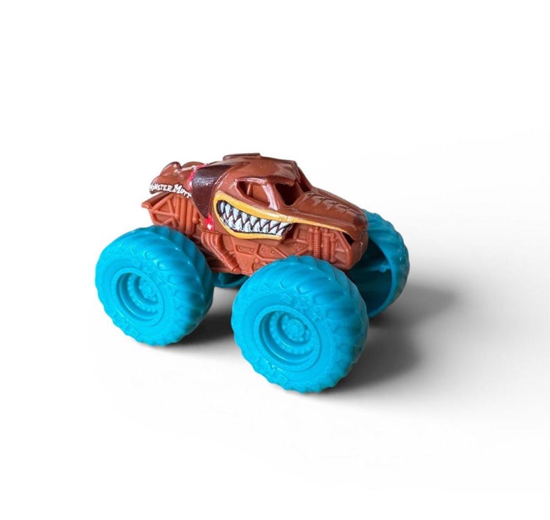 K&ouml;nnte beinhalten: Ein braunes Monstertruck-Spielzeug mit detailliertem Design, mit einem grinsenden Mund und t&uuml;rkisblauen Reifen. Das Spielzeug hat die Aufschrift "Monster Mutt" an der Seite. Das Spielzeug steht vor einem wei&szlig;en Hintergrund.