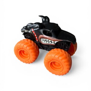 K&ouml;nnte beinhalten: Schwarzes Monstertruck-Spielzeug mit leuchtend orangefarbenen R&auml;dern. Der Truck hat ein hunde&auml;hnliches Gesichtsdesign mit scharfen Z&auml;hnen und roten Details. Die gro&szlig;en orangefarbenen R&auml;der sind f&uuml;r das Gel&auml;nde geeignet.