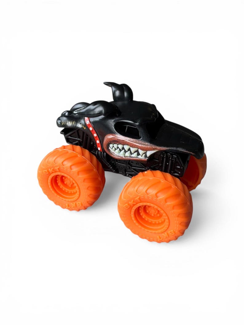K&ouml;nnte beinhalten: Schwarzes Monstertruck-Spielzeug mit leuchtend orangefarbenen R&auml;dern. Der Truck hat ein hunde&auml;hnliches Gesichtsdesign mit scharfen Z&auml;hnen und roten Details. Die gro&szlig;en orangefarbenen R&auml;der sind f&uuml;r das Gel&auml;nde geeignet.