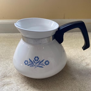 May include: White ceramic coffee pot with a black handle and a silver metal band. The pot features a blue floral design. The coffee pot is designed with a spout for easy pouring.
