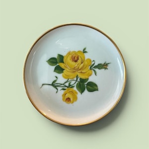 May include: A white ceramic plate with a gold rim, featuring a yellow rose design with green leaves. The plate is round and has a delicate floral pattern, suitable for serving or display.
