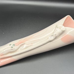 May include: A white ceramic vase with a pink interior and exterior accents. The vase has a unique, folded design with a sculpted floral element and green accents. The vase is approximately 12 inches long.