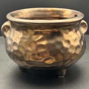 May include: A bronze-colored, hammered-texture ceramic pot with three small feet and two side handles. The small decorative bowl has a glossy finish and is suitable for holding small items or as a decorative accent piece.