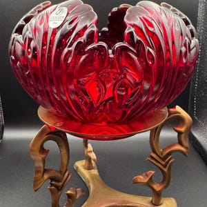 May include: A ruby red Fenton glass bowl with a floral design, resting on a decorative brass stand. The bowl has a textured surface and is handmade in the USA. The stand has an ornate, three-legged design.