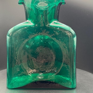 May include: A vibrant green glass vase with a square base and a rounded top. The vase has a circular design in the center and a label with text. The vase is made by Blenko Handcraft.