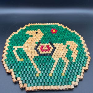 May include: Round, woven coaster with a green background and a central design featuring two light brown figures and a red and black center. A small red flower is in the upper right corner. The coaster is made of small, cylindrical pieces.