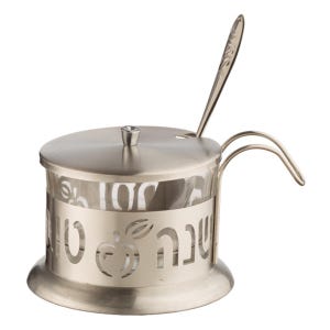 May include: A silver-toned sugar bowl with a lid and a small spoon. The bowl has a clear glass interior and a decorative band with Hebrew text and an apple design. The handle is curved, and the lid has a small crystal-like knob.