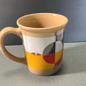 May include: A tan ceramic mug featuring a graphic design with a large circular motif divided into four quadrants of black, white, red, and yellow. The design includes thin vertical lines and small horizontal dashes on a white and yellow background.