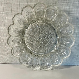 May include: Clear glass egg serving dish with a scalloped edge. The dish has a central circular area and 12 egg-shaped indentations around the perimeter. The glass has a textured, dotted pattern.