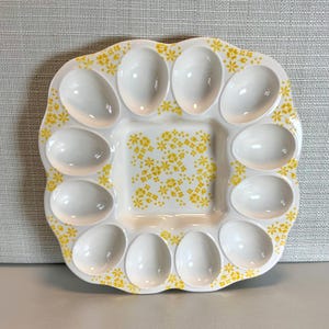 May include: White ceramic deviled egg serving platter with 12 egg-shaped indentations around a square center. The platter is decorated with yellow floral patterns. A decorative serving dish for appetizers.