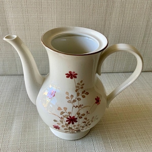 May include: Cream-colored ceramic teapot with a floral design. The teapot features a curved spout, a rounded body, and a handle. The floral pattern includes red flowers and brown leaves. The rim and handle are trimmed with a brown accent.