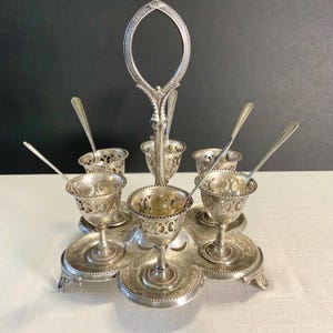 May include: A silver-colored egg cup stand with six egg cups and spoons. The stand has a central handle and a circular base with ornate detailing. Each egg cup has a decorative, openwork design.