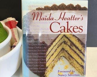 Puede incluir: Un libro de cocina de tapa dura titulado Maida Heatter's Cakes con un prólogo de Nancy Silverton. La portada presenta una gran imagen de un pastel amarillo de varias capas con glaseado de chocolate oscuro y una lista de recetas de pasteles.