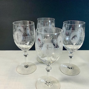 May include: Four clear wine glasses with etched designs. Each glass features a central oval with a figure holding a bow and arrow, surrounded by decorative patterns. The glasses have a round base and a detailed stem.
