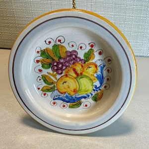 May include: A round, ceramic plate with a hand-painted fruit design. The plate features a yellow rim, a light blue border, and a brown inner rim. The center of the plate displays a colorful arrangement of grapes, apples, and other fruits.