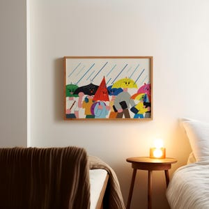 May include: A framed art print depicts a colorful illustration of people with umbrellas in the rain. The artwork features figures with various umbrella colors, including yellow, green, and pink, under blue rain lines. The frame is made of light-colored wood.