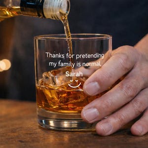 May include: A clear glass tumbler filled with ice and amber liquid, with more liquid being poured from a bottle. The glass has the text "Thanks for pretending my family is normal, Sarah" and a smiley face.