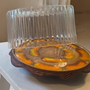 May include: A round ceramic serving dish with a brown, wood-textured rim and a glossy center featuring a pattern of orange and yellow curved shapes, topped with a clear, vertically ribbed, cylindrical plastic cover for food protection.