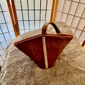 May include: A brown leather tote bag with a stitched white seam down the front. The bag has a single handle and a triangular shape. The bag is sitting on a gray, textured surface.