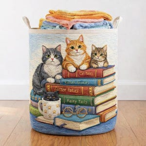 May include: A blue and beige tapestry laundry hamper featuring three cats sitting on a stack of books. The books have titles like "Cat Tales" and "Fairy Tails." The hamper is filled with folded towels and has two handles.