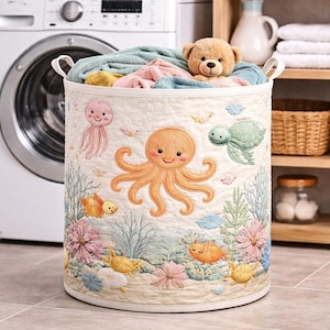Könnte beinhalten: Ein runder, cremefarbener Wäschekorb mit einem gesteppten Design mit einem Oktopus, einer Qualle, einer Schildkröte und Fischen. Der Korb ist mit gefalteten Handtüchern und einem Teddybären gefüllt. Das Design umfasst Korallen- und Blumenelemente.