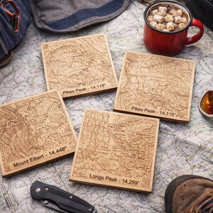 May include: Four square wooden coasters with engraved topographic maps of mountain peaks, including Pikes Peak and Mount Elbert. The coasters are surrounded by outdoor gear, including a backpack, sunglasses, and a mug of hot chocolate.