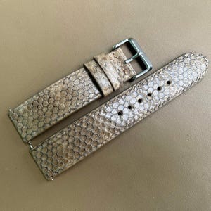 Può includere: Un cinturino per orologio in due pezzi con una trama testurizzata a pelle di serpente nei toni del beige e del marrone chiaro. Il cinturino include una fibbia in metallo argentato e due passanti abbinati, con fori di regolazione circolari.