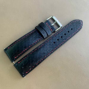 May include: A black watch strap with a textured, scale-like pattern and contrasting red stitching. The strap features a silver-toned metal buckle and two matching black leather keepers, designed for secure attachment to a watch case.