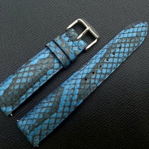 Puede incluir: Una correa de reloj de dos piezas con un patrón texturizado de piel de serpiente en azul y negro. La correa incluye una hebilla de metal plateado pulido y dos trabillas a juego en el segmento corto, con agujeros perforados en el segmento largo.