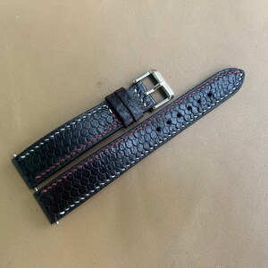 May include: A black leather watch strap featuring a textured honeycomb pattern, accented with contrasting red and white stitching. The strap includes a silver-toned metal buckle and a matching leather keeper loop, designed for a standard wristwatch.