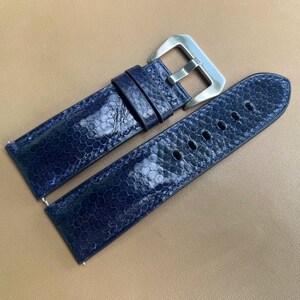 Può includere: Un cinturino per orologio blu scuro caratterizzato da un distinto motivo a scaglie esagonali sulla superficie. Il cinturino include una fibbia in metallo color argento lucido e due passanti abbinati, con una serie di fori per la regolazione.