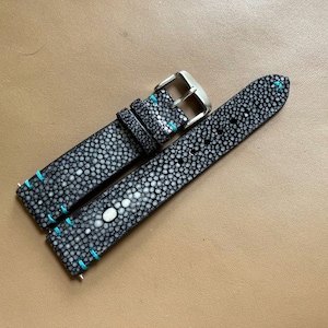 May include: A black stingray leather watch strap featuring a textured, pebbled surface with a prominent white central eye pattern. The strap includes bright turquoise stitching along the edges and a polished silver-tone metal buckle and loops.