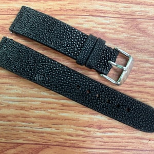 May include: A black watch strap featuring a textured, pebbled surface finish. The strap includes a silver-toned metal buckle and a matching sliding keeper loop, designed for attachment to a timepiece with a standard spring bar mechanism.