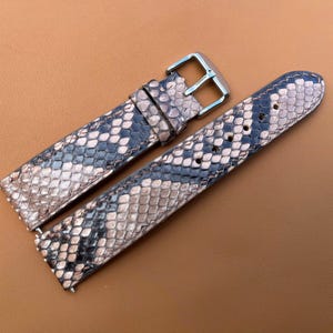 May include: A two-piece watch strap featuring a textured snakeskin pattern in shades of beige, gray, and black. The strap includes a silver-toned metal buckle and a matching sliding keeper loop, with pre-punched holes for adjustable sizing.