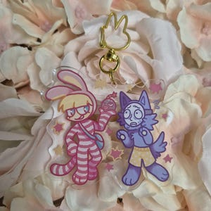 以下が含まれることがあります： 金色のウサギ型の留め金が付いた、2つの漫画のキャラクターキーホルダー。1つのキーホルダーは、小さなオブジェクトを持っているピンクと白のストライプのウサギのキャラクターが特徴です。もう1つのキーホルダーは、黄色のショーツを着た紫色の猫のようなキャラクターが特徴です。