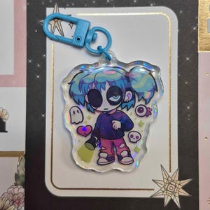 Könnte beinhalten: Ein holografischer Anhänger mit einer Cartoonfigur mit blauen Haaren und einer Maske, die eine Taschenlampe hält. Der Anhänger hat einen blauen Verschluss und Grafiken eines Geistes, Herzens, Auges und Schädels. Der Hintergrund ist eine dekorative Karte.