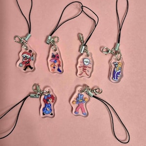 Puede incluir: Siete colgantes de acrílico transparente con diseños de personajes de dibujos animados. Cada colgante tiene un personaje diferente en varios colores, sujeto a un cordón negro con un cierre plateado. Los colgantes se muestran sobre un fondo rosa.