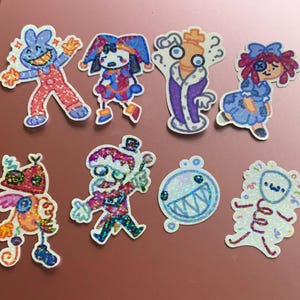 May include: A collection of eight colorful, glittery stickers featuring whimsical cartoon characters. Designs include a jester, a doll, a king, and other unique figures. The stickers have a white border and are set against a reddish-brown background.