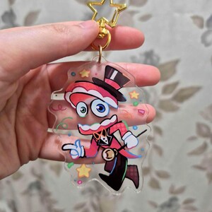 May include: A clear acrylic keychain featuring a cartoon magician character in a red and black outfit, top hat, and star-shaped gold clasp. The character has large blue eyes and is pointing with one hand. Colorful confetti and stars surround the character.