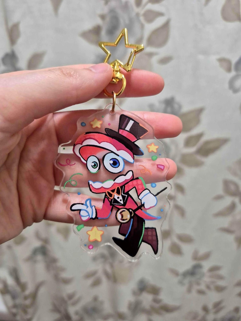 May include: A clear acrylic keychain featuring a cartoon magician character in a red and black outfit, top hat, and star-shaped gold clasp. The character has large blue eyes and is pointing with one hand. Colorful confetti and stars surround the character.