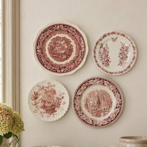 May include: Four decorative ceramic plates with maroon transferware patterns featuring pastoral landscapes and floral motifs are mounted on a wall above a wooden surface holding two stacked books, a small bowl, and two cream-colored pitchers.