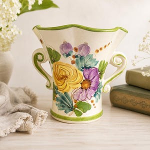 May include: A cream-colored ceramic vase with a scalloped rim and two scrolled handles. The exterior features hand-painted floral motifs in shades of yellow, purple, and green, with a green painted band around the base and the rim edge.
