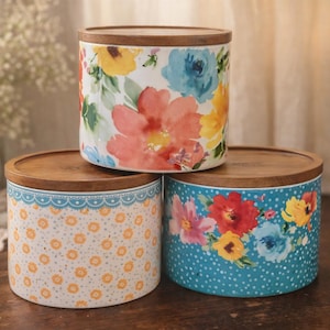 May include: Three ceramic canisters with wooden lids. The canisters feature floral patterns in various colors, including red, yellow, and blue. One canister has a blue and white dotted pattern. The canisters are stacked.