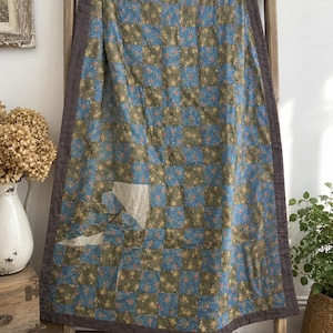 May include: A rectangular patchwork quilt featuring a grid of alternating blue and olive green floral fabric squares. The quilt is bordered by a solid dark gray fabric and includes a visible area of light-colored fabric damage on the lower left side.